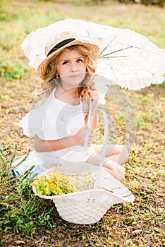 A nice little girl with light curls in a straw hat, a white dress and a lace umbrella with a basket of flowers in the summer field