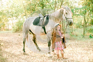 A nice little girl with light curly hair in a vintage plaid dress and a straw hat and a gray horse