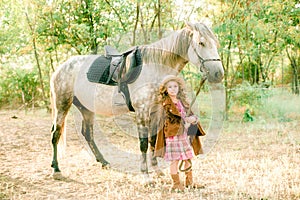A nice little girl with light curly hair in a vintage plaid dress and a straw hat and a gray horse