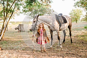 A nice little girl with light curly hair in a vintage plaid dress and a straw hat and a gray horse