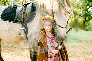 A nice little girl with light curly hair in a vintage plaid dress and a straw hat and a gray horse