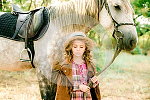 A nice little girl with light curly hair in a vintage plaid dress and a straw hat and a gray horse