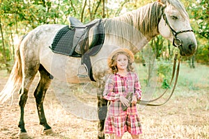 A nice little girl with light curly hair in a vintage plaid dress and a straw hat and a gray horse