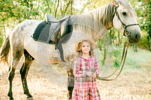 A nice little girl with light curly hair in a vintage plaid dress and a straw hat and a gray horse