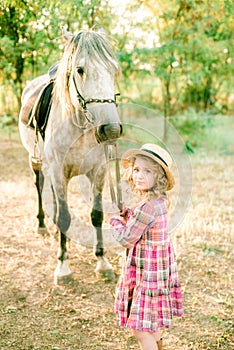 A nice little girl with light curly hair in a vintage plaid dress and a straw hat and a gray horse