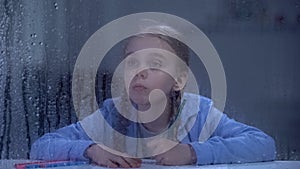 Nice little girl painting behind rainy window, orphan child dreaming about home