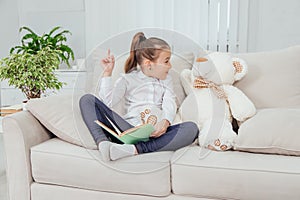 Nice little girl sitting on the sofa with teddy-bear near her, reading fairy-tale, pointing her finger up, saying photo
