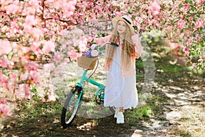 Nice longhaired blonde girl in pink jacket and straw hat walking in blooming apple garden with retro bicycle