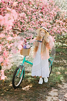 Nice longhaired blonde girl in pink jacket and straw hat walking in blooming apple garden with retro bicycle and smiling