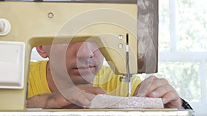 A nice man is sitting at the electric sewing machine at home. He sews several layers of fabric and serves a working tool