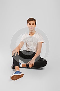 Nice man in white t-shirt, black trousers and sneakers half laying on the floor over grey background