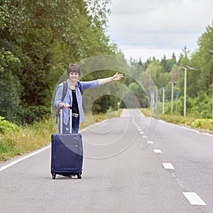 Nice middle-aged woman with a travel bag hitchhiking on the rural road in summer