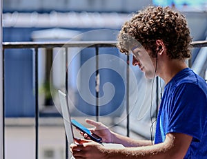 Nice photo of a young Caucasian man working with his laptop, earphones and mobile phone. He is sitting on the attic window,