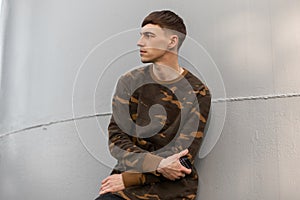 Nice serious young man in a stylish green military shirt c fashionable hairstyle rests near the metal gray wall