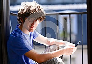 Nice shot of a young Caucasian man with his cell phone. He is sitting on an attic window, he looks into the camera smiling.