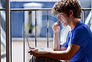 Nice shot of a young Caucasian man working with his laptop talking on earphones. He is sitting on an attic window, facing the sun