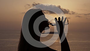 Nice silhouette of unrecognizable girl drink water from transparent bottle at sunset, in background ocean or sea with