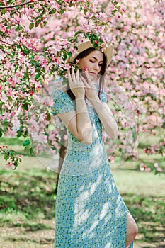 Nice slim young girl in blue dress and straw hat stay in blooming  garden