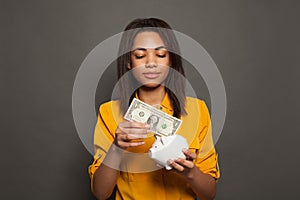 Nice smart black woman student putting in piggy bank one dollar banknote