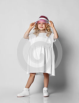 Nice smiling blonde curly kid girl princess in white casual dress and sneakers is standing holding her cap