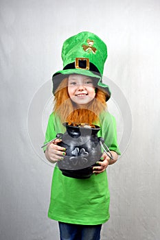 Nice smiling small girl with decorative red beard, green clover leaf on her cheek and leprechaun hat, holds cast iron pot, full of