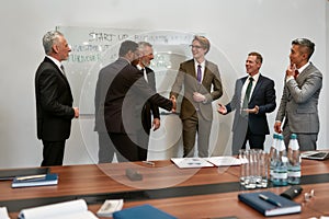 Nice to meet you. Two happy businessmen in classic wear shaking hands while having a meeting with their team in the