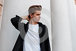 Nice trendy young man in a stylish black shirt in a white t-shirt with a fashionable hairstyle stands near the white columns
