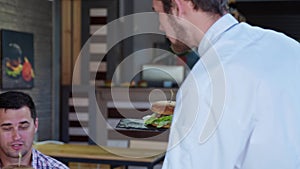 Nice waiter makes tasty juicy burgers to joyful and hungry visitors of happy family with children having fun in cafe in