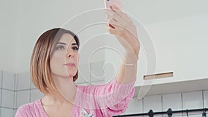 A nice woman is holding a smartphone, making selfie in the kitchen.