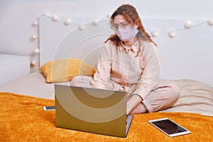 A nice woman in a medical mask is sitting on a home bed and remotely working on a laptop. A girl in a face mask works at a