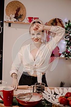 A nice, young blonde girl alone in a white shirt and jeans in the  kitchen. The kitchen is decorated with New Year`s decor. New Ye