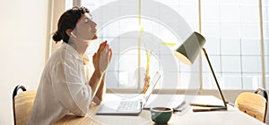 Nice young caucasian girl is praying with palms together and closing eyes indoors by window.