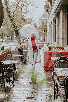 Nice young girl in white coat and red sweater walking with umbrella under the rain