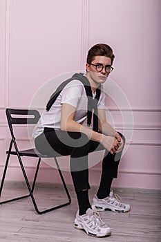 Nice young man model in glasses in fashionable youth black-white clothes in trendy leather sneakers posing sitting on a vintage