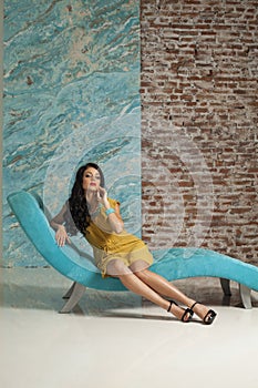 Nice young woman make-up and long hair wearing yellow dress posing sitting on blue trendy comfortable sofa