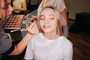 Nice young woman sit in beauty room with closed eyes. She smile. Make up artist put eyeshadows on eyeline. She hold photo