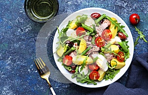 Nicoise salad with canned tuna, tomatoes, boiled eggs, green beans, potatoes and olives on white plate, blue table background,