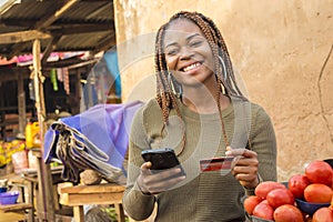 nigerian lady selling in a local nigerian market using her mobile phone and credit card to do a transaction online smiling