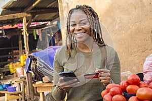 nigerian woman selling in a local nigerian market using her mobile phone and credit card to do a transaction online smiling