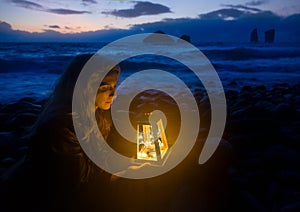 Night at the beach, blonde woman with lantern, waves of the sea and wild rock formations in the background