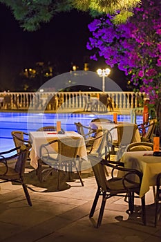 Night cafe, empty tables served for dinner, candles, lights, by the pool in the garden with palm trees and flowers, evening rest