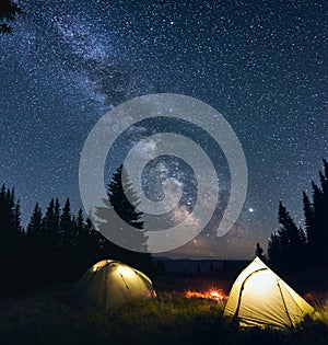 Night camp with burning bonfire and two tents in pine forest under bright starry sky on which the Milky Way