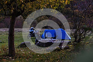 Night camp on the forest. Camping with big blue tent and touring motorcycle, Woman is sitting on folding chairs use smatphone.