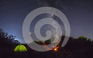 Night camping. Couple tourists have a rest at a campfire near illuminated tent under amazing night sky