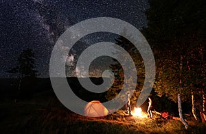 Night camping. Guy and girl standing by fire near woods and tents under starry sky