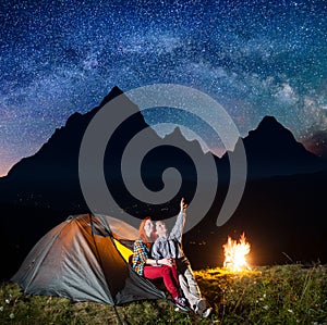 Night camping. Happy couple tourists sitting near tent and fire and enjoying incredibly beautiful starry sky, Milky way