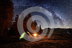 Night camping. Illuminated tent and campfire near forest under night sky full of stars and milky way