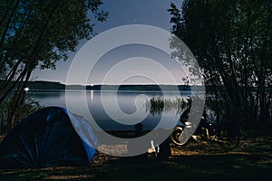 Night camping on lake shore. Man and woman is sitting. Couple tourists enjoying amazing view of night sky full of stars. Blue tent