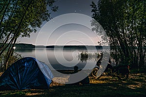 Night camping on lake shore. Man and woman is sitting. Couple tourists enjoying amazing view of night sky full of stars. Blue tent photo