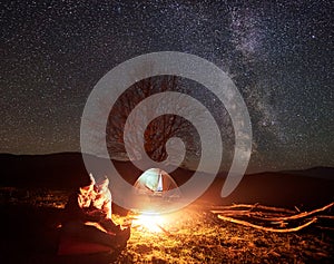 Night camping in mountains. Couple hikers having rest near campfire, tourist tent under starry sky